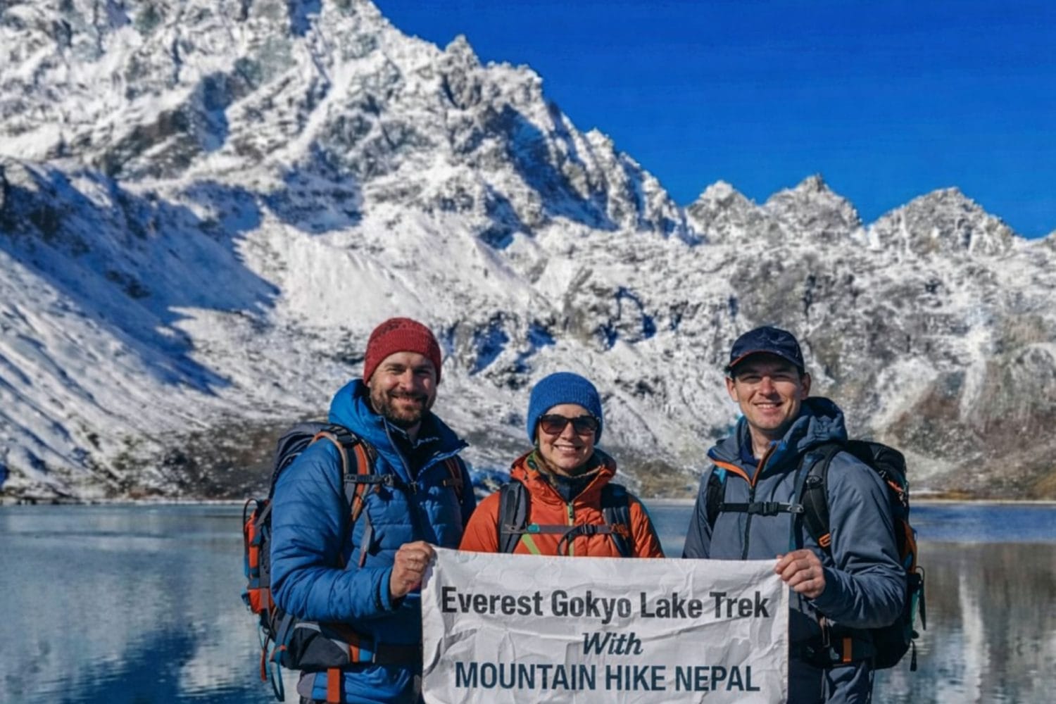 Trekkers at Gokyo Lake holding Everest Gokyo Lakes Trek banner with snow-covered Himalayan peaks