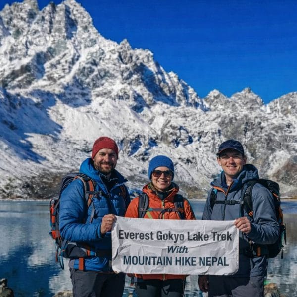 Trekkers at Gokyo Lake holding Everest Gokyo Lakes Trek banner with snow-covered Himalayan peaks