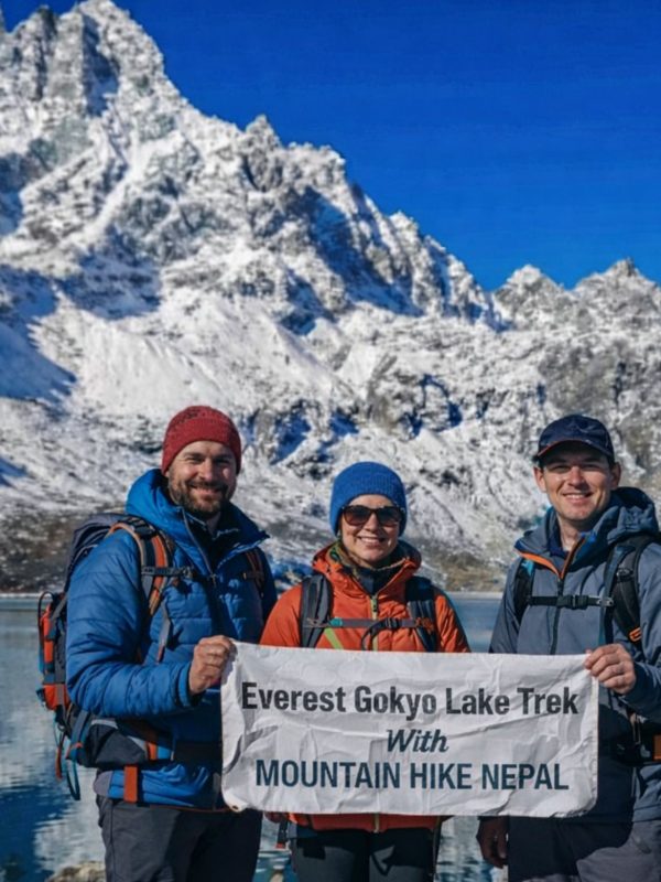 Trekkers at Gokyo Lake holding Everest Gokyo Lakes Trek banner with snow-covered Himalayan peaks