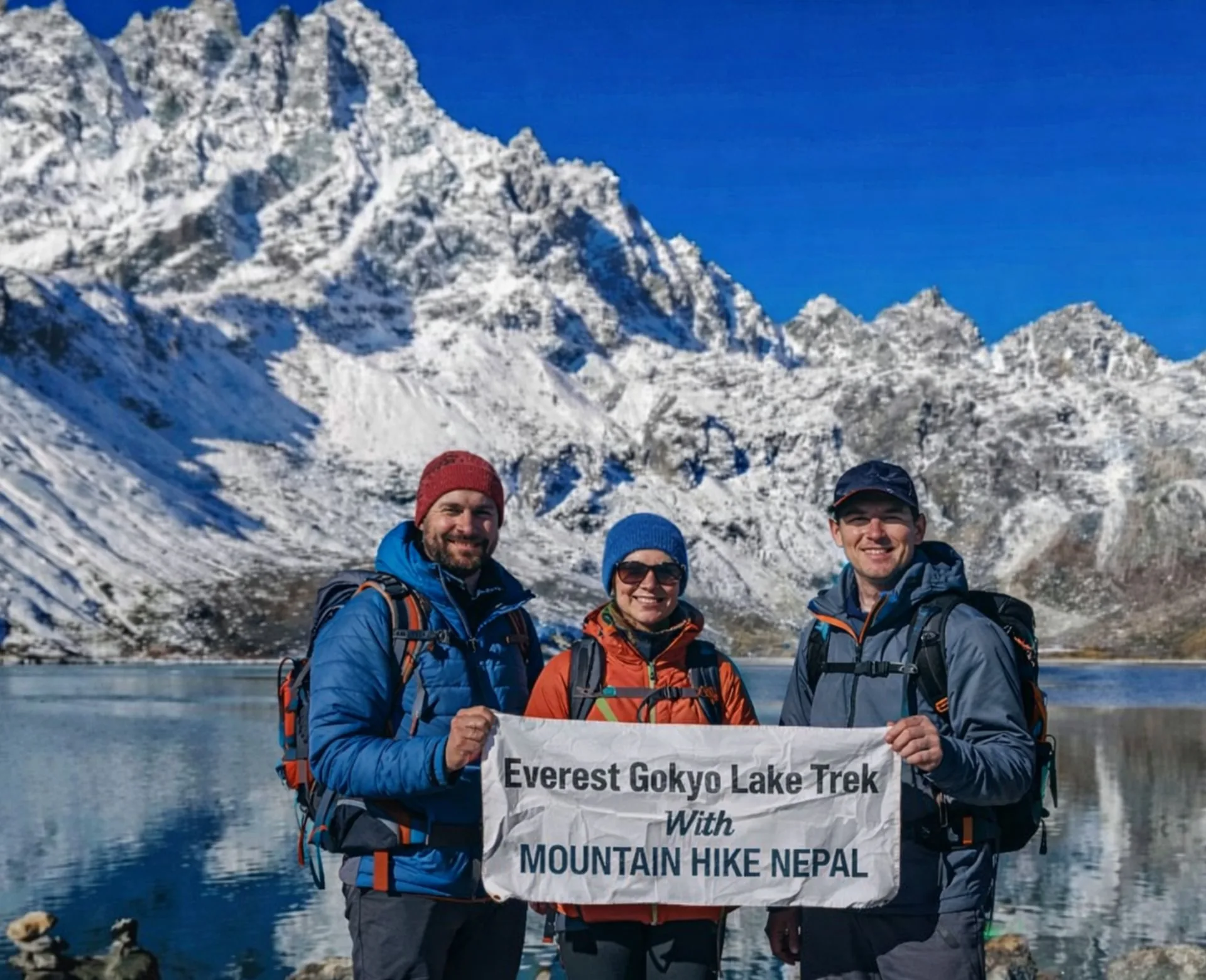 Trekkers at Gokyo Lake holding Everest Gokyo Lakes Trek banner with snow-covered Himalayan peaks