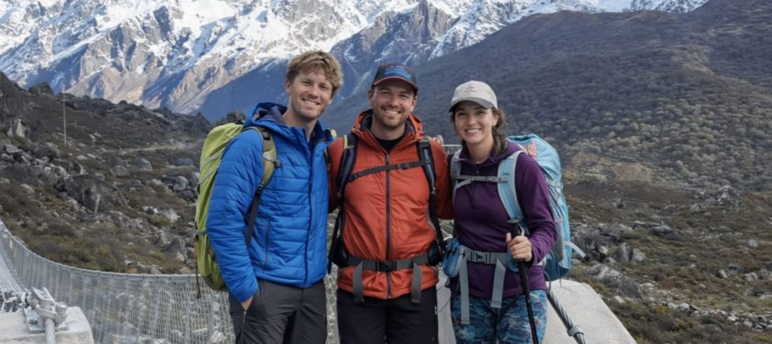 Three trekkers with backpacks crossing a suspension bridge with snow-capped Himalayan peaks in the background, Nepal