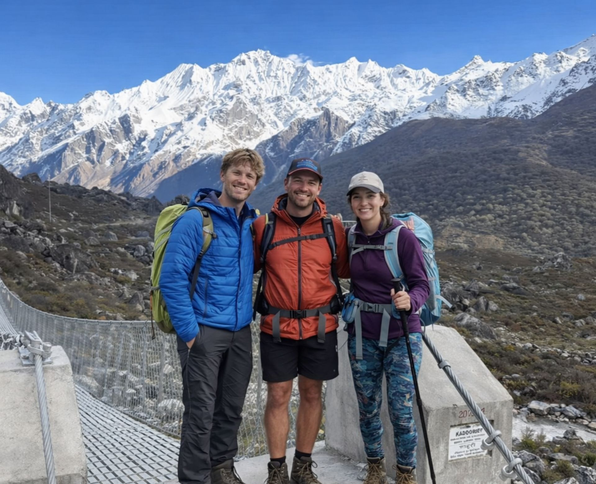 Three trekkers with backpacks crossing a suspension bridge with snow-capped Himalayan peaks in the background, Nepal