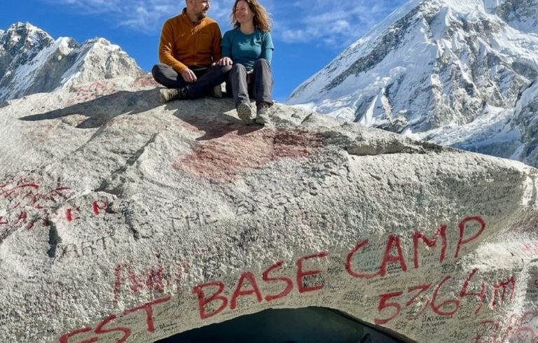 Beginner trekkers sitting at the Everest Base Camp marker surrounded by Himalayan peaks during the Everest Base Camp Trek in Nepal.