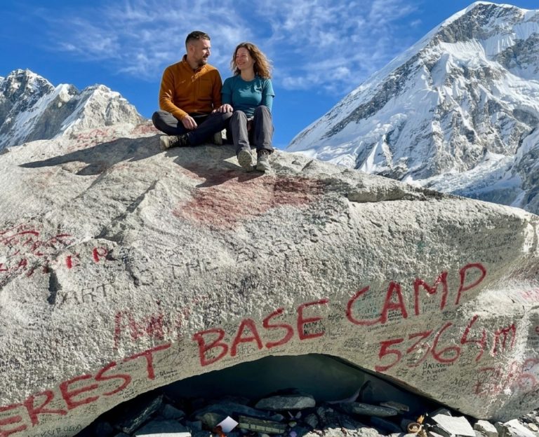 Beginner trekkers sitting at the Everest Base Camp marker surrounded by Himalayan peaks during the Everest Base Camp Trek in Nepal.