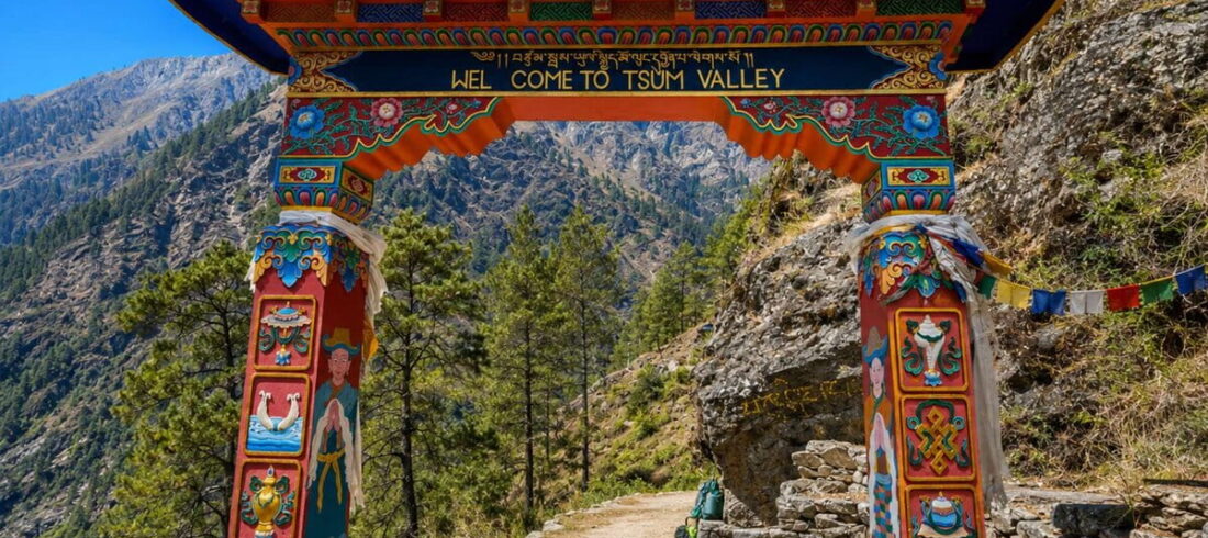 Painted Buddhist gate with a “Welcome to Tsum Valley” sign over a stone trail, with pine forest and steep hills behind