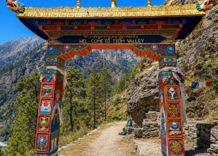 Painted Buddhist gate with a “Welcome to Tsum Valley” sign over a stone trail, with pine forest and steep hills behind