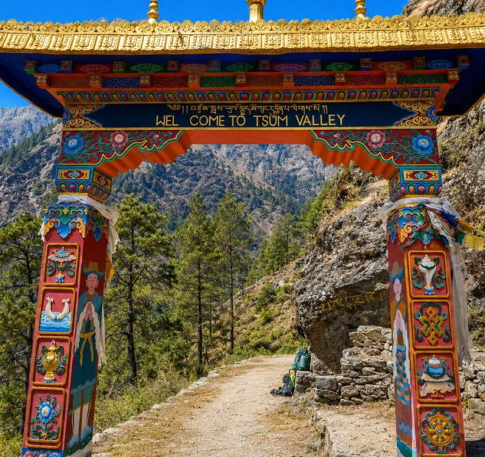 Painted Buddhist gate with a “Welcome to Tsum Valley” sign over a stone trail, with pine forest and steep hills behind