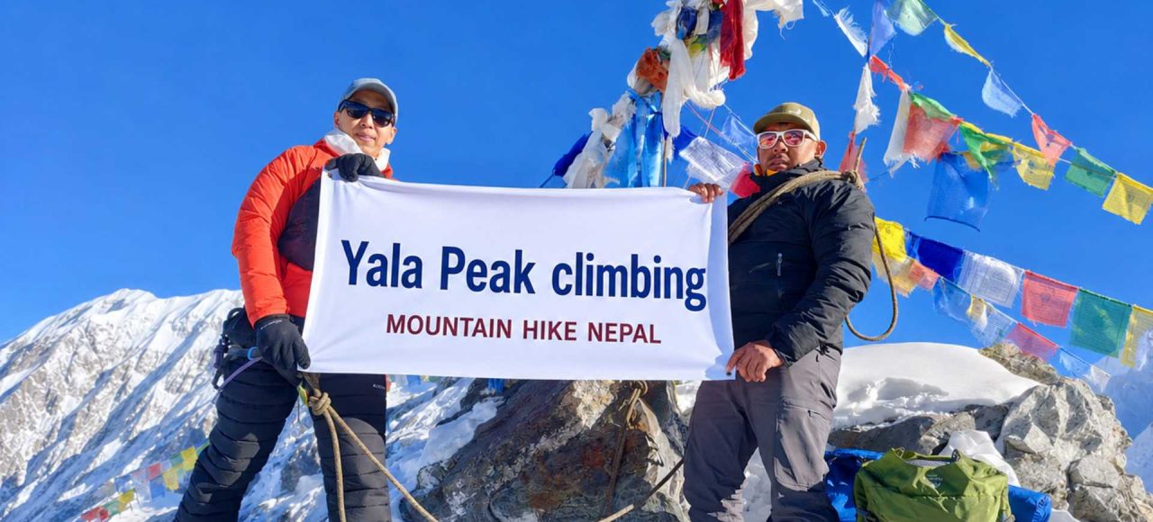 Climbers holding a Yala Peak Climbing banner at the 5,732m summit in Langtang Valley, Nepal with snow-covered Himalayan mountains and prayer flags in the background.