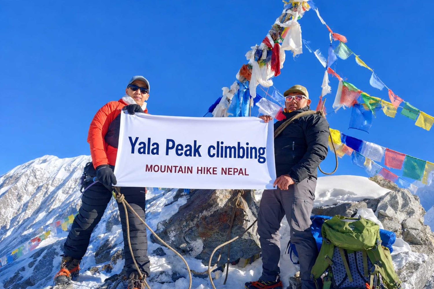 Climbers holding a Yala Peak Climbing banner at the 5,732m summit in Langtang Valley, Nepal with snow-covered Himalayan mountains and prayer flags in the background.