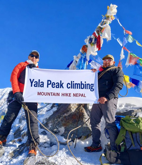 Climbers holding a Yala Peak Climbing banner at the 5,732m summit in Langtang Valley, Nepal with snow-covered Himalayan mountains and prayer flags in the background.