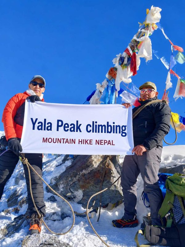 Climbers holding a Yala Peak Climbing banner at the 5,732m summit in Langtang Valley, Nepal with snow-covered Himalayan mountains and prayer flags in the background.