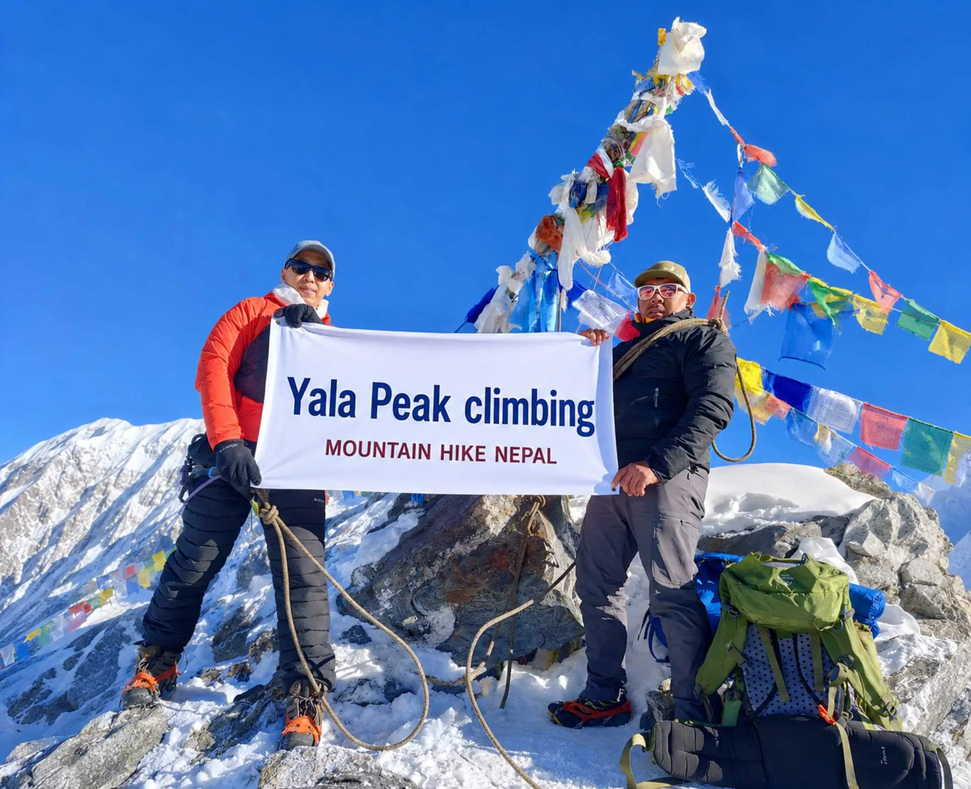 Climbers holding a Yala Peak Climbing banner at the 5,732m summit in Langtang Valley, Nepal with snow-covered Himalayan mountains and prayer flags in the background.
