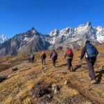 A group of hikers with backpacks and trekking poles walking along an alpine ridge trail toward snow-capped Himalayan peaks in Sagarmatha National Park, Nepal.