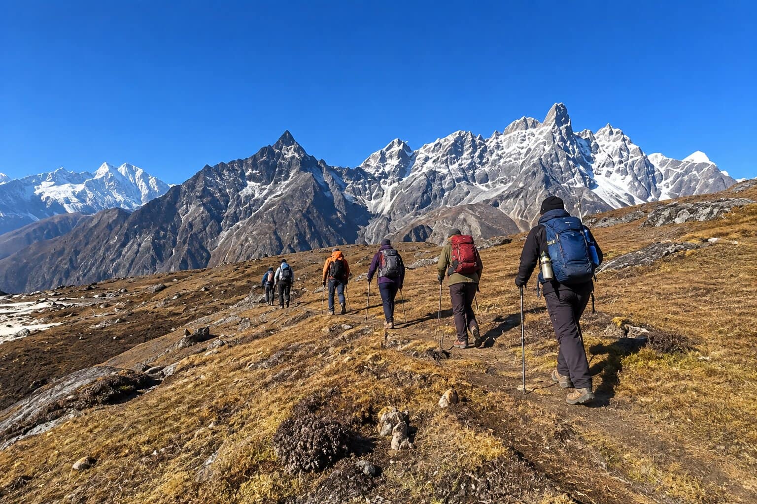 A group of hikers with backpacks and trekking poles walking along an alpine ridge trail toward snow-capped Himalayan peaks in Sagarmatha National Park, Nepal.