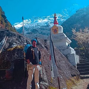 Trekker with a backpack and trekking poles beside a white stupa, with snow-covered peaks behind, in the Everest region.